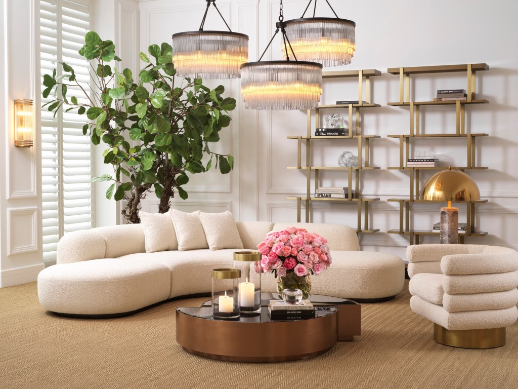 A stylish living room featuring a cream bouclé sofa, a round glass coffee table with candles and a floral arrangement, a plant, elegant chandeliers, and modern shelving.