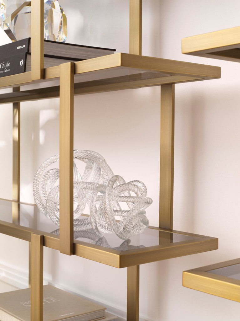 A close-up of a decorative glass sculpture resembling intertwined loops, displayed on a stylish gold metal shelving unit with books and crystal accents.