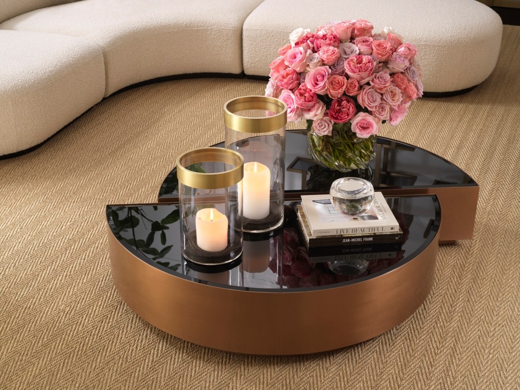 A stylish coffee table with a half-moon design featuring a glossy black surface, a bouquet of pink roses, two glass candle holders with gold rims, and a small decorative item on top of stacked books.
